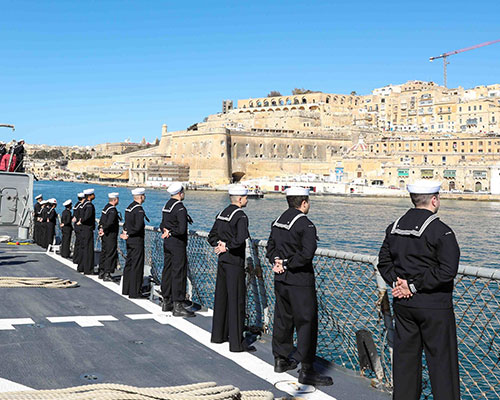Sailors standing at attention on the side of a ship pulling into a European port.